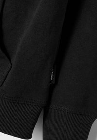 Felpa nera con una trama di cotone morbido, polsini a coste e un'etichetta laterale. Il design è semplice e privo di ornamenti, evidenziando la funzionalità.