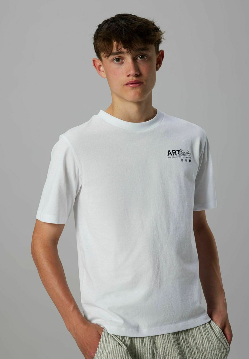 Jonge man met kort bruin haar, gekleed in een wit T-shirt met de tekst "ART Studio" op de borst en gestreepte broek, poserend tegen een grijze achtergrond.