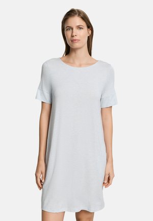 Kobieta o prostych brązowych włosach, ubrana w jasnoniebieską, codzienną sukienkę z krótkim rękawem, sięgającą kolan, stojąca na tle jednolitej białej ściany.