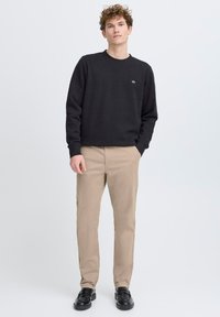 Felpa nera con collo a giro e logo ricamato, abbinata a pantaloni beige e scarpe nere, indossata da una persona posizionata al centro.