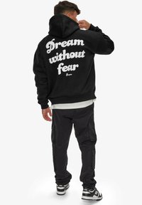 Hoodie noir avec le texte "Rêve sans peur" en blanc dans le dos, associé à un pantalon cargo noir et des baskets noir et blanc.