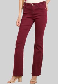 Donna che indossa pantaloni bordeaux a vita alta e gamba dritta con un dettaglio della vita intrecciato e sandali beige con tacco, in piedi davanti a uno sfondo neutro.