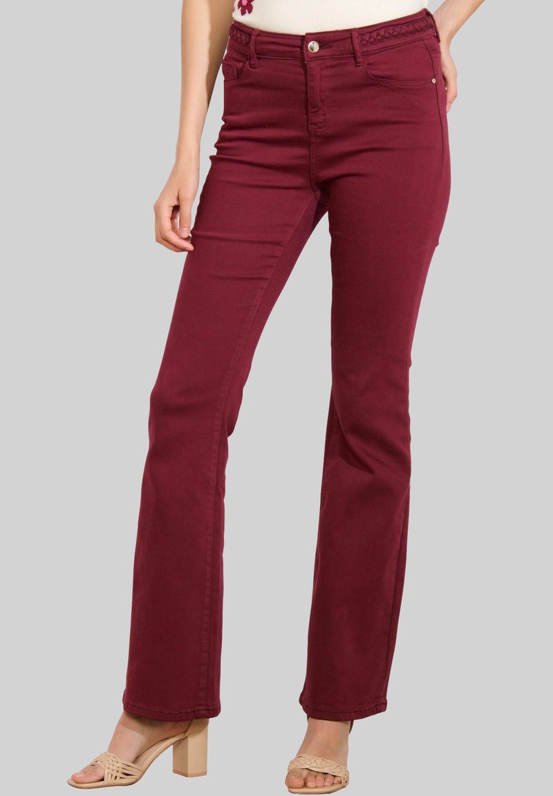Donna che indossa pantaloni bordeaux a vita alta e gamba dritta con un dettaglio della vita intrecciato e sandali beige con tacco, in piedi davanti a uno sfondo neutro.
