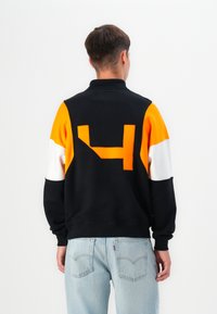 Mustekala ja oranssi värilohkottu huppari, jonka selässä on suuri oranssi numero "4", ja kontrastoivat valkoiset ja oranssit hihat.