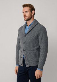 Cárdigan de punto acanalado gris con cuello de chal, con cierres de botón y bolsillos frontales, llevado sobre una camisa azul. Material texturizado.