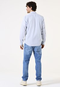 Lichtblauwe, langemouwige overhemd met knopen en een gestructureerde stof, gedragen met vervaagde blauwe jeans en beige sneakers, gezien vanuit de achterkant.
