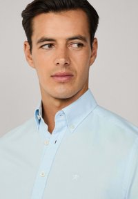 Camicia azzurra chiara con una texture morbida; presenta un piccolo logo bianco sul lato sinistro del petto e un colletto standard con dettaglio a bottoni.
