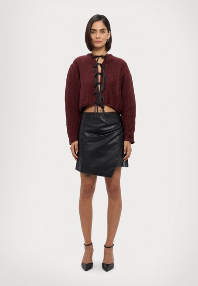 Burgundijas krāsas īsais džemperis ar melniem aukliņu akcentiem, savienots ar melnu ādas apvalka svārku, izceļot gludas tekstūras un piegulošu siluetu.