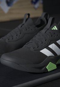 Paire de chaussures de sport noires avec des rayures blanches et des accents verts reposant sur une surface sombre.