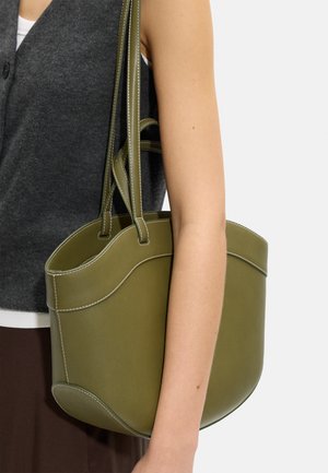 Kobieta nosząca ciemny top bez rękawów trzyma oliwkowozieloną skórzaną torebkę z podwójnymi paskami na ramieniu na jednolitym tle.