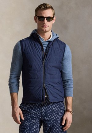 Man met donkere zonnebril, marineblauw doorgestikt vest over blauwe trui en gemêleerde marineblauwe broek tegen een effen achtergrond.