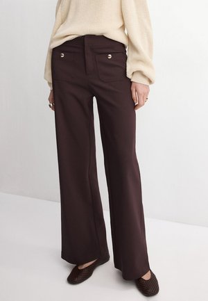 Persoană purtând pantaloni largi maro închis cu detalii cu nasturi aurii pe buzunarele din spate, un pulover tricotat crem și pantofi slip-on împletiți de culoare închisă.