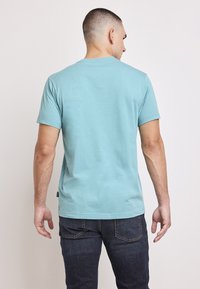 Ljusblå bomullströja med korta ärmar och rund halsringning, bärs av en person sedd bakifrån, kombinerad med mörka jeans.