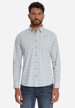 Man draagt een wit overhemd met lange mouwen en een klein geometrisch patroon en een blauwe spijkerbroek, staand tegen een effen achtergrond.