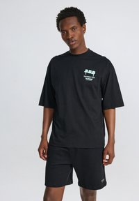 Svart bomullströja med korta ärmar, med en grön grafisk design på vänster bröst. Matchas med svarta shorts i minimalistisk stil.