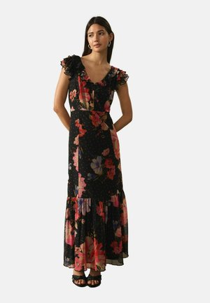 Moteris vilki juodą gėlėtą maksi suknelę su raukšlėtais trumpais rankovėliais ir sandalais, stovi su rankomis už nugaros.