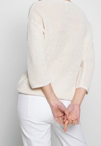 Jasno beżowy sweter dzianinowy z rękawami o długości trzech czwartek, o luźnym kroju i prążkowanymi mankietami, w połączeniu z białymi spodniami.