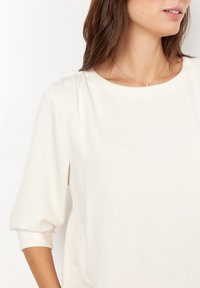 Mujer con una blusa de manga larga color blanco roto y cuello redondo, adornada con un delicado collar de oro con un pequeño colgante.
