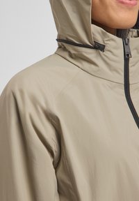 Beige waterdichte jas met een gestroomlijnd ontwerp, zwarte rits en capuchon. De stof heeft een gladde, licht glanzende textuur.