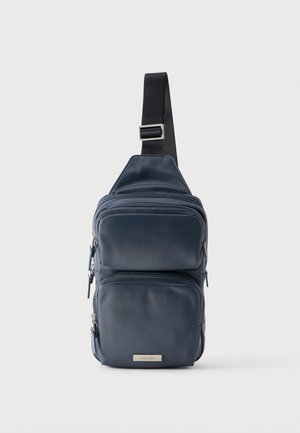 Sac bandoulière bleu marine avec sangle noire réglable, trois compartiments zippés et petite plaque métallique logo en bas à l'avant.