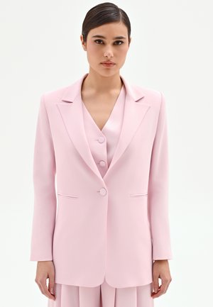 Donna con capelli scuri che indossa un blazer rosa pallido su misura, sopra un gilet abbottonato coordinato e una gonna plissettata, su uno sfondo chiaro uniforme.