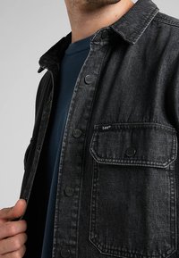 Černá denimová košile s knoflíkovým zapínáním vpředu, náprsní kapsou a značkovými knoflíky. Nosená přes modrou košili s klasickým límcem a strukturovanou tkaninou.