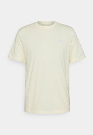 Kort cremet Nike T-shirt med et lille hvidt broderet logo på venstre bryst, vist mod en ensfarvet baggrund.