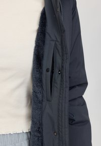 Dark navy puffer jacket with a fluffy interior lining, featuring snap button pockets and a smooth exterior fabric. Zipper closure at front.