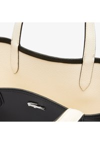 Sac fourre-tout texturé crème avec garniture noire et poignées, avec poche intérieure noire ornée d'un logo crocodile blanc.