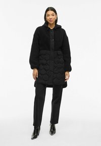 Melns quiltots mētelis ar mīkstu, teksturētu augšdaļu. Iespējo kapuci, aizdari ar spraudēm un divas priekšējās kabatas. Nēsāts virs melnām biksēm un zoli augstiem zābakiem.