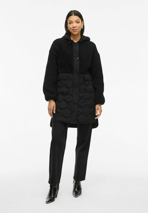 Schwarze gesteppte Jacke mit plüschigem, strukturiertem Obermaterial. Verfügt über eine Kapuze, Druckknopfverschluss und zwei Vorderaschen. Trägt sie über schwarzen Hosen und Absatzstiefeln.