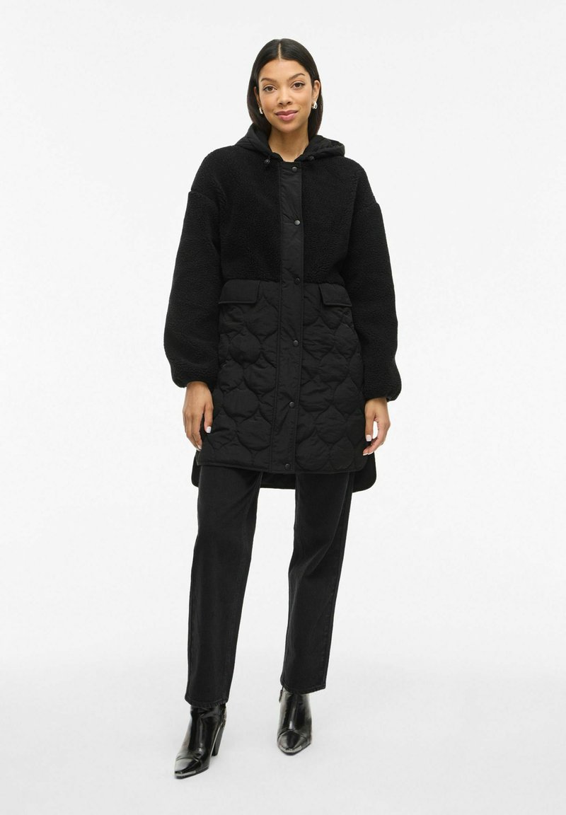 Melns quiltots mētelis ar mīkstu, teksturētu augšdaļu. Iespējo kapuci, aizdari ar spraudēm un divas priekšējās kabatas. Nēsāts virs melnām biksēm un zoli augstiem zābakiem.