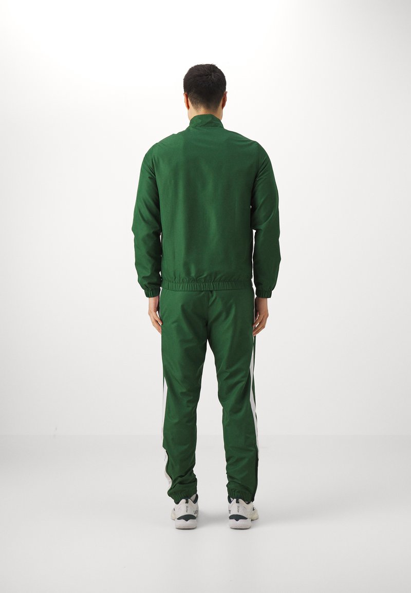 Homme de dos portant un survêtement vert foncé avec des rayures blanches sur les côtés et des sneakers blanches sur un fond blanc uni.