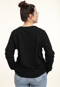 Felpa nera a collo rotondo con maniche lunghe, polsini e orlo a coste, realizzata in tessuto morbido. Indossata con jeans azzurri, mostrata da dietro.
