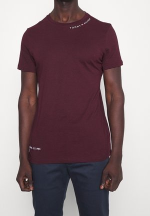 Mann trägt ein eng anliegendes bordeauxrotes Tommy Hilfiger T-Shirt mit weißem Logo-Schriftzug nahe am Kragen und dunkelblaue Hose.
