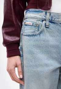 Κοντινό πλάνο ενός χεριού που ακουμπά δίπλα στην τσέπη ανοιχτομπλε τζιν Calvin Klein, με ένα μανίκι μαρον δερμάτινου μπουφάν ορατό.