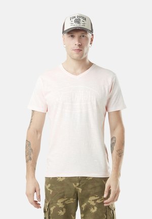 Homme portant des boucles d'oreilles, une casquette beige Von Dutch, un t-shirt rose clair à col en V et un short cargo camouflage, debout devant un fond uni.