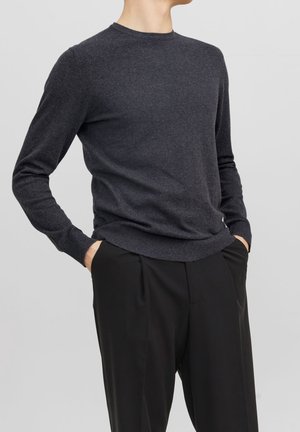 Dunkelgrauer Rundhals-Pullover aus weichem Strickmaterial, mit langen Ärmeln und geripptem Saum. Getragen mit schwarzen Hosen, die Hände in den Taschen.