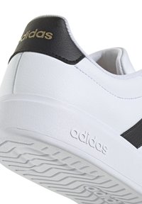 Zapatilla blanca con detalles en negro, parte superior sintética texturizada, suela plana de goma y logo en relieve en el lateral.