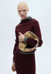 Burgunderfarbener Strickpullover mit grauen Akzenten, kombiniert mit einer beigen und braunen Handtasche mit Muster, die einen geschlungenen Griff und metallische Details aufweist.