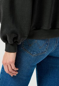 Czarna bluza z marszczonymi rękawami w połączeniu z niebieskimi dżinsami, które mają charakterystyczny detal w postaci V-kształtnego przeszycia na kieszeniach.