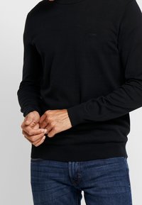Black knit sweater with a high neckline, made from smooth material. Features long sleeves and a textured ribbed hem. Subtle logo on chest.