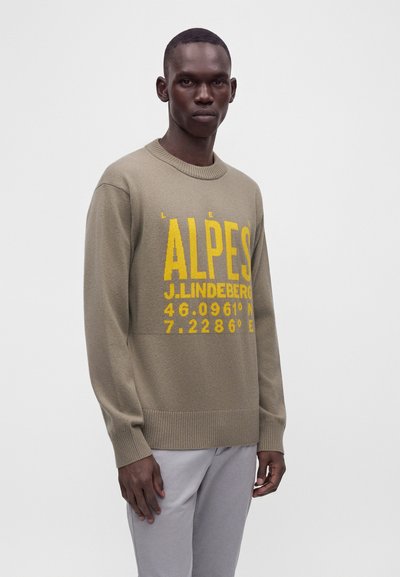 Homme portant un pull beige avec un texte jaune "ALPES J. LINDEBERG" et des coordonnées, associé à un pantalon gris clair, debout devant un fond uni.