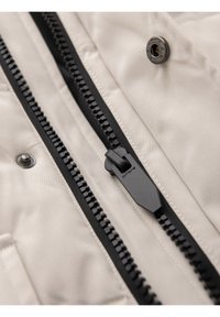 Gros plan d'une fermeture éclair noire et d'un bouton-pression métallique sur un tissu beige clair d'une veste ou d'un manteau.
