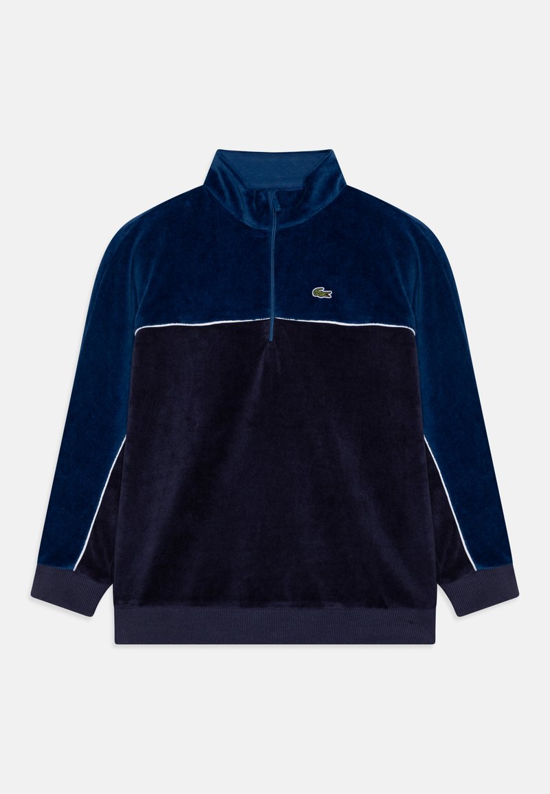 Pull en polaire bleu marine avec une bande d'accent blanche. Il présente un col montant et une fermeture éclair sur le devant à quart. Logo vert sur la poitrine gauche.