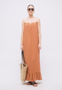 Kvinde iført stropløs maxikjole i rustfarve, sorte sandaler, solbriller og holder en stor flettet shopper med sorte håndtag.