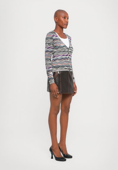 Cardigan cu un model zigzag multicolor, cu decolteu în V, asortat cu o fustă mini din piele maro, cu fermoar lateral. Pantofi cu toc înalt negri.