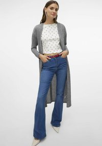 Grå lång kofta över en vit topp med röd blommönster, tillsammans med blå utsvängda jeans och ett rött bälte. Vita spetsiga klackar.