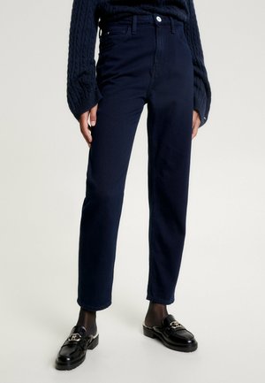 Femme portant un pantalon droit taille haute bleu marine foncé, des mocassins noirs avec des ornements dorés et un pull en maille torsadée bleu marine.