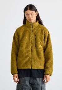 Veste en polaire de couleur vert olive avec fermeture éclair intégrale, col haut et poche poitrine zippée. Tissu texturé et doux, coupe décontractée.